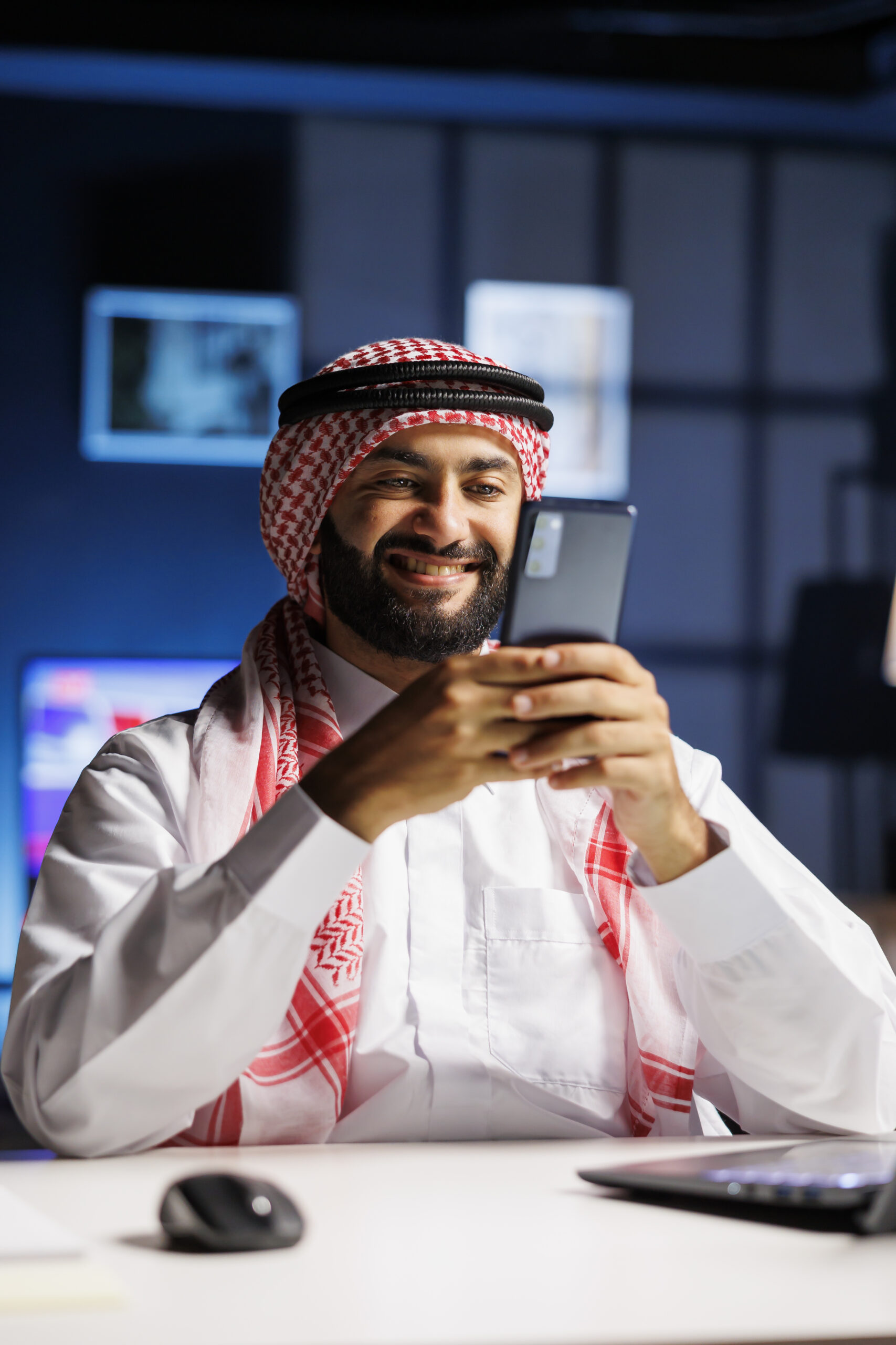 Detailed view of youthful man in Arabian clothing using his cell phone for online communication. Healthy Muslim guy with a smile, browsing the internet on his modern smartphone.